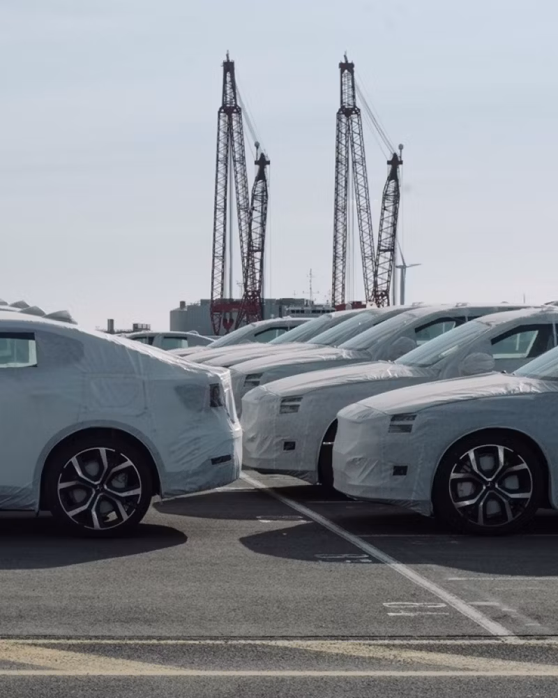 Polestar cars covered in protective wrap parked in rows near cranes and wind turbine in an industrial area.